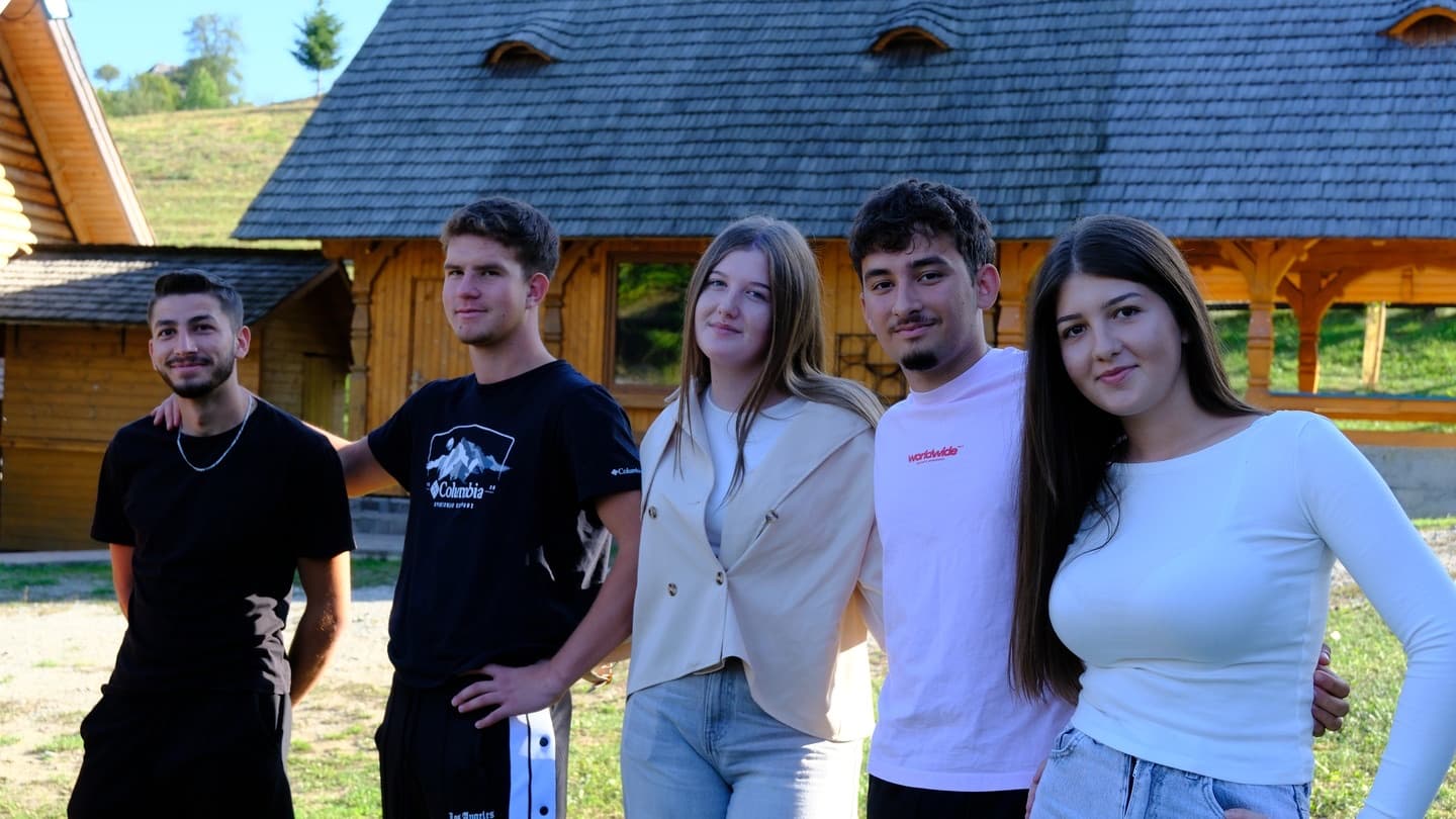 Cinci tineri zâmbind în fața unei case de lemn